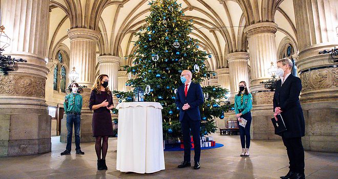 Das Friedenslicht vor dem Weihnachtsbaum in der Rathaus Diele, daneben stehen Carola Veit, Dr. Peter Tschentscher und die Bischöfin Kirsten Fehrs, sowie ein Pfadfinder und eine Pfadfinderin.