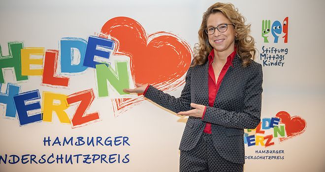 Carola Veit vor einer Wand mit dem Logo des Hamburger Kinderschutzpreises „Heldenherz