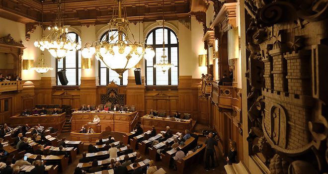 Blick in den Plenarsaal, in dem eine Bürgerschaftssitzung stattfindet. Rechts im Vordergrund ein geschnitztes Hamburg-Wappen an der Wand.