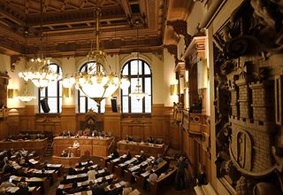 Blick in den Plenarsaal, in dem eine Bürgerschaftssitzung stattfindet. Rechts im Vordergrund ein geschnitztes Hamburg-Wappen an der Wand.