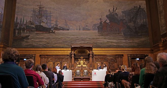 Froschperspektive zur Bühne aus der Mitte des Ganges zwischen den Stuhlreihen des Publikums, oberhalb der Debattant:innen ein großes Gemälde.
