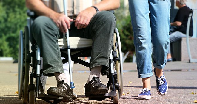 Ein älterer Mann in einem Rollstuhl, neben ihm läuft eine junge Frau.