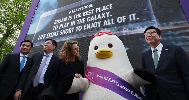 Blick von unten zu vier Personen auf der Bühne vor einem großen Bildschirm mit einem lebensgroßen pinguinähnlichen Maskottchen für die Expo 2030 in Busan.