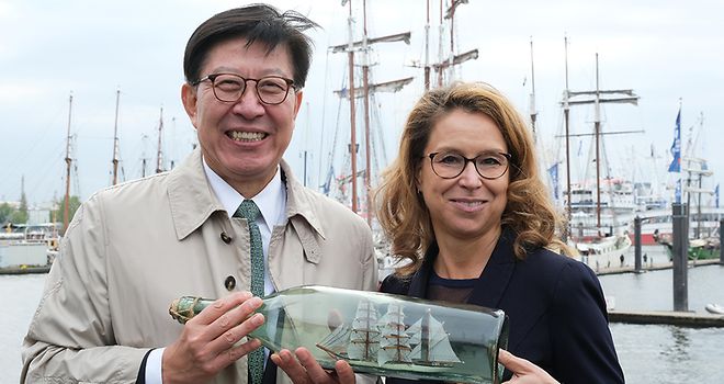 Busans Bürgermeister Park Heong-joon und Präsidentin Carola Veit blicken in die Kamera und halten ein Buddelschiff, im Hintergrund sind Masten von Segelschiffen zu sehen.