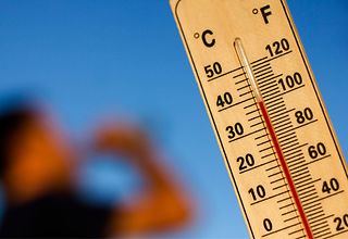 Ein Thermometer zeigt Temperaturen um die 38 Grad Celsuis an. In Hintergrund trinkt ein Menschen aus einer Flasche.