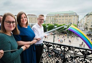 Mareike Engels, Katharina Fegebank und Christoph Kahrmann stehen auf dem Rathausbalkon und halten die Kordel mit der Regenbogenflagge in der Hand.