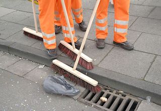 Einige Personen in orangen Arbeitshosen mit Besen bei der Reinigung einer Straße.