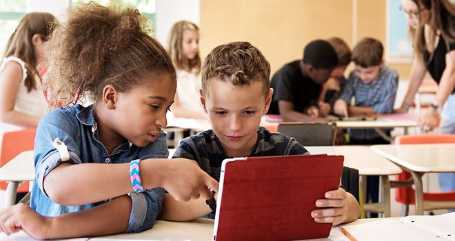 Ein Mädchen und ein Junge sitzen in einem Klassenraum und schauen gemeinsam auf ein Tablet..