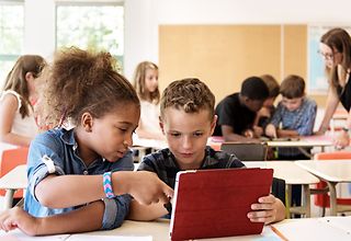 Ein Mädchen und ein Junge sitzen in einem Klassenraum und schauen gemeinsam auf ein Tablet..