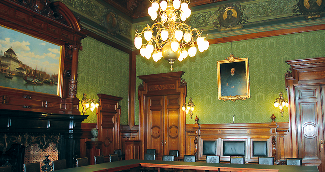 Mittig im Bürgersaal stehen Tische in rechteckiger Formation. Außen herum stehen Stühle. Der Raum ist mit einer prunkvollen grünen Tapete ausgestattet. An den Wänden hängen historische Gemälde.