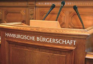 Auf dem Rednerpult steht der Schriftzug "Hamburgischen Bürgerschaft" und es sind drei Mikrofone auf dem Pult befestigt.