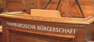 Auf dem Rednerpult steht der Schriftzug "Hamburgischen Bürgerschaft" und es sind drei Mikrofone auf dem Pult befestigt.