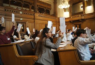 Jugendliche im Plenarsaal stimmen bei einer Sitzung von „Jugend im Parlament“ ab. Dazu halten sie rote Zettel nach oben.