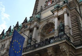 Blick von unten nach oben zum Rathausbalkon an dem die Flagge der EU und die Hamburger Flagge wehen.
